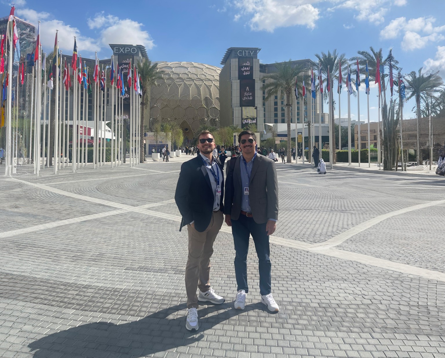 Two attendees outside Expo City in Dubai, with national flags and the Al Wasl dome behind them